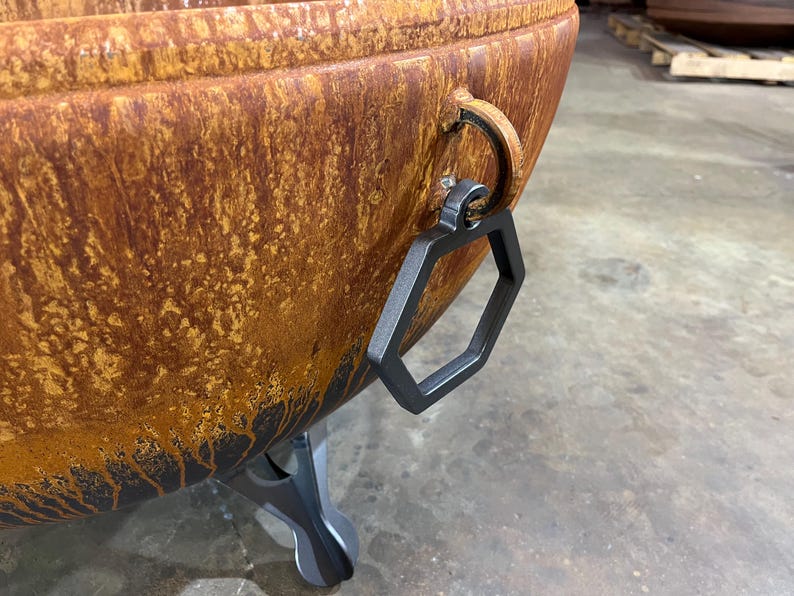 May include: A large, bowl-shaped fire pit with a weathered, rusted orange and brown patina. It features a matte black hexagonal metal handle attached to a circular ring on the side and is supported by a sturdy, dark gray metal tripod base.