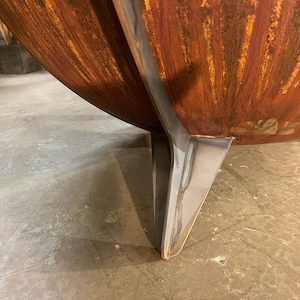 Puede incluir: Un brasero en forma de cuenco con un exterior de metal oxidado de color naranja con un acabado texturizado. El recipiente est&aacute; sostenido por una pata robusta de acero inoxidable de color plateado liso que se extiende por el lado curvo.