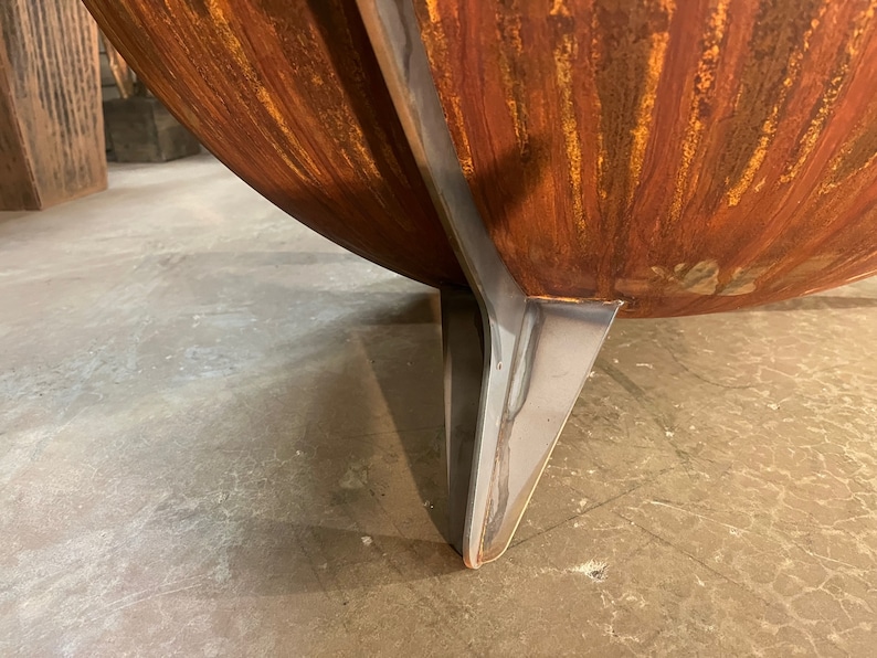 Puede incluir: Un brasero en forma de cuenco con un exterior de metal oxidado de color naranja con un acabado texturizado. El recipiente est&aacute; sostenido por una pata robusta de acero inoxidable de color plateado liso que se extiende por el lado curvo.