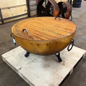 May include: A large, circular, rust-colored metal fire pit bowl featuring a matching flat lid with two arched handles. The bowl is supported by three sturdy, dark-colored metal legs and includes additional metal ring handles on the sides.