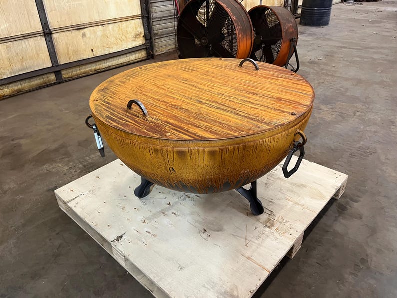 May include: A large, circular, rust-colored metal fire pit bowl featuring a matching flat lid with two arched handles. The bowl is supported by three sturdy, dark-colored metal legs and includes additional metal ring handles on the sides.