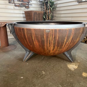 Puede incluir: Una gran hoguera de metal en forma de cuenco con un acabado de p&aacute;tina oxidada en tonos naranja y marr&oacute;n. Presenta un borde de metal gris oscuro liso, tres patas anguladas resistentes y una peque&ntilde;a placa de metal ovalada con texto.