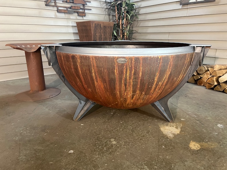 Puede incluir: Una gran hoguera de metal en forma de cuenco con un acabado de p&aacute;tina oxidada en tonos naranja y marr&oacute;n. Presenta un borde de metal gris oscuro liso, tres patas anguladas resistentes y una peque&ntilde;a placa de metal ovalada con texto.