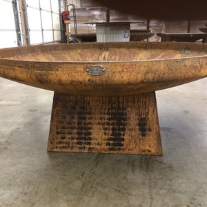 May include: A large, rusty metal fire pit with a round bowl and a square base. The fire pit has a metal tag with the word "Wazari" on it.