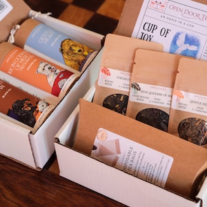 Puede incluir: Una caja blanca abierta con una tapa marrón está llena de diferentes bolsitas de té y tubos de té. Las bolsitas de té están etiquetadas con los nombres de los tés, como "Iron Goddess of Mercy" y "Gentle Heart". Los tubos de té están etiquetados con los nombres de los tés, como "Coconut" y "Iron Goddess of Mercy". La caja está etiquetada con "Open Door Tea" y "An Oasis in a Cup".