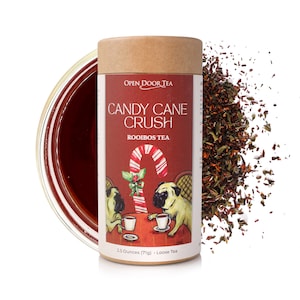 Puede incluir: Un envase cilíndrico de té rooibos "Candy Cane Crush". La etiqueta presenta una ilustración de bastón de caramelo y dos perros pug. El envase está junto a un montón de hojas de té sueltas y un vaso de té preparado. Contiene 71g de té suelto.