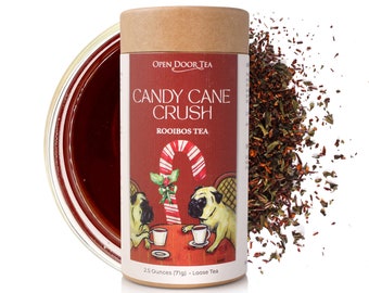 May include: A cylindrical container of "Candy Cane Crush" rooibos tea. The label features a candy cane illustration and two pug dogs. The container is next to a pile of loose tea leaves and a glass of brewed tea. The container holds 2.5 ounces (71g) of loose tea.