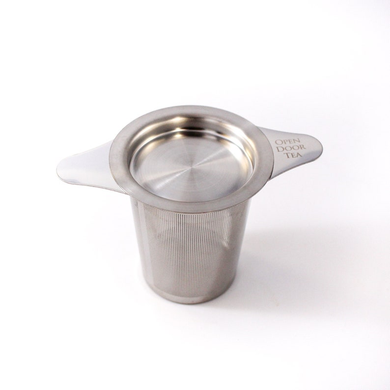 May include: A silver tea infuser with a lid and a handle. The handle has the text "OPEN DOOR TEA" engraved on it.