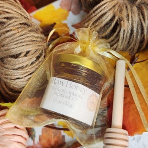 May include: A jar of raw honey, labeled "Raw Honey, Wermuth Apiaries, Stratford, CT, 15 oz (42 g)", is presented in a gold organza bag. A wooden honey dipper and twine are also visible. The background features autumn leaves.
