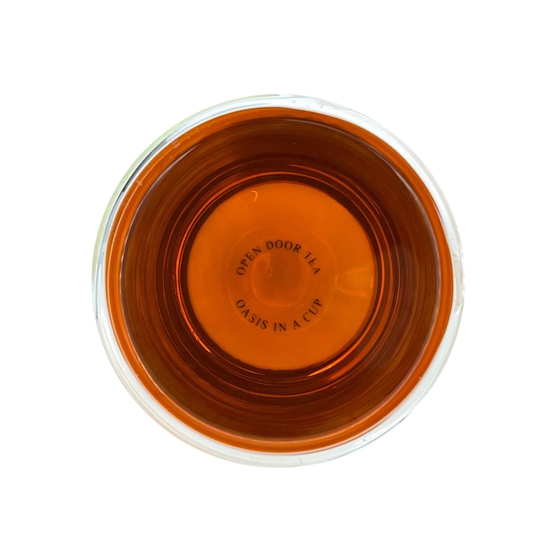 May include: A clear glass cup filled with amber-colored tea. The bottom of the cup has the text "Open Door Tea Oasis in a Cup" printed on it.