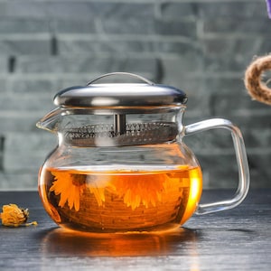 May include: A clear glass teapot with a stainless steel lid and infuser, filled with orange tea and flower blossoms. The teapot has a curved handle and a spout for pouring. The tea is a warm, inviting color.