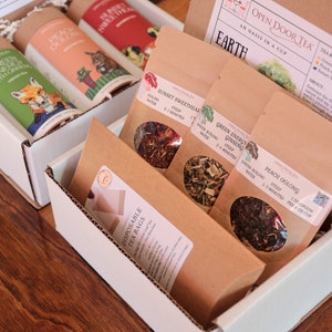 May include: A white box with a brown cardboard insert containing six individual tea bags. The tea bags are labeled with the names "Green Energy Ginseng", "Peach Oolong", "Sunset Sweetheart", and "Green Tea". The box is labeled "Open Door Tea: An Oasis in a Cup".