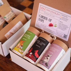 May include: A white box with four brown cardboard tubes containing loose leaf tea. The tubes are labeled with the names of the teas: Jasmine Dragon, Earl Grey Rose, and Serenity Rose. The box is labeled "Open Door Tea" and "Garden of Hope".