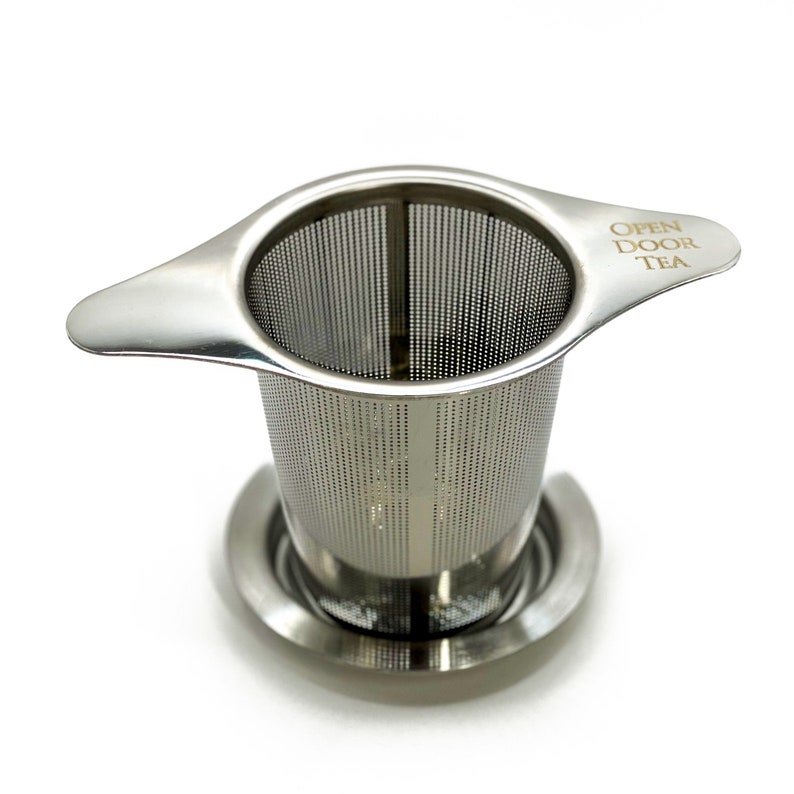 May include: A stainless steel tea infuser with a handle and a mesh filter. The infuser has a base and the text "OPEN DOOR TEA" is engraved on the handle.