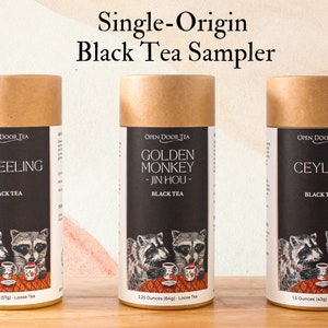 Könnte beinhalten: Ein Sampler mit drei schwarzen Tees in zylindrischen Behältern. Die Behälter haben einen braunen Deckel und ein schwarzes Etikett mit dem Namen des Tees und einer Waschbär-Illustration. Der Text "Single-Origin Black Tea Sampler" steht oben. Die Tees sind Darjeeling, Golden Monkey und Ceylon.
