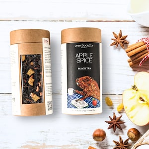 May include: Two cylindrical tea containers with brown lids. One displays tea leaves with fruit pieces. The other features "Apple Spice Black Tea" with a beaver illustration. Cinnamon sticks, star anise, and apple slices surround the tea.