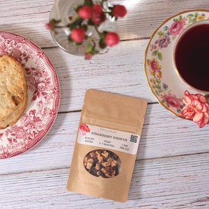 May include: A brown paper pouch with a clear window showing strawberry hibiscus tea inside. The pouch has text that reads "Strawberry Hibiscus" and instructions for brewing the tea. The tea is loose leaf and includes dried fruit pieces.