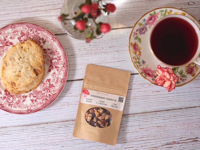 May include: A brown paper pouch with a clear window showing strawberry hibiscus tea inside. The pouch has text that reads "Strawberry Hibiscus" and instructions for brewing the tea. The tea is loose leaf and includes dried fruit pieces.