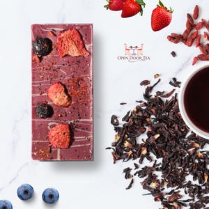 Op de afbeelding: Een rechthoekige reep donkere chocolade met gedroogd fruit en blauwe bessen. De chocolade is versierd met witte swirls. De reep ligt op een wit marmeren oppervlak. Op de achtergrond is een kopje thee, gedroogde hibiscusbloemen en gedroogde gojibessen te zien. De tekst "Open Door Tea" is zichtbaar op de afbeelding.