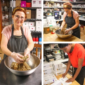 May include: Three people working in a tea shop. A woman in a pink shirt and black and white striped apron mixes loose leaf tea in a silver bowl. Another woman in a white shirt and black and white striped apron mixes loose leaf tea in a silver bowl. A person in a red shirt is organizing boxes of tea.