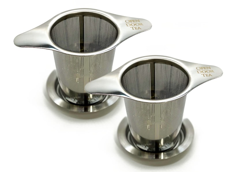 May include: Two stainless steel tea infusers with handles and a base. The infusers have a fine mesh and the handles are engraved with the words "Open Door Tea".