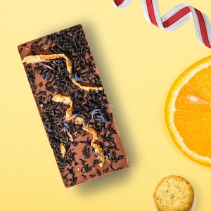 May include: A rectangular chocolate bar with tea leaves, orange peel, and blue flower petals on a yellow background. A slice of orange, a cookie, and a red and white ribbon are also present.