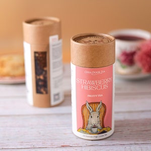 May include: Two cardboard tubes of loose leaf tea. The tube on the left is brown with white text. The tube on the right is brown with a pink label that says "Strawberry Hibiscus Fruity Tea" and features a cartoon rabbit sitting in a chair.
