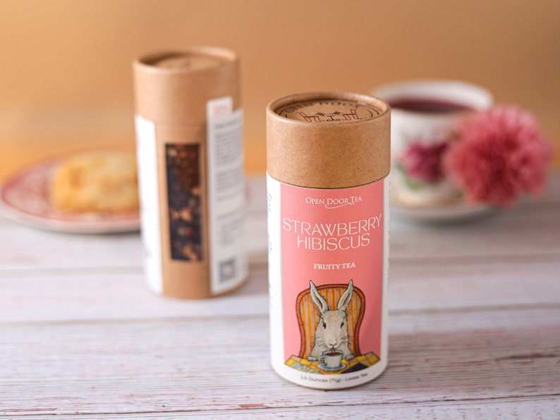 May include: Two cardboard tubes of loose leaf tea. The tube on the left is brown with white text. The tube on the right is brown with a pink label that says "Strawberry Hibiscus Fruity Tea" and features a cartoon rabbit sitting in a chair.