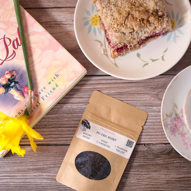 May include: A brown paper pouch of loose leaf Pu-Erh Berry tea with a clear window showing the dark tea leaves. The pouch has text that reads "Open Doorlia Pu-Erh Berry Boiling Water Steep 3-5 Minutes 1 Teaspoon Per Cup".