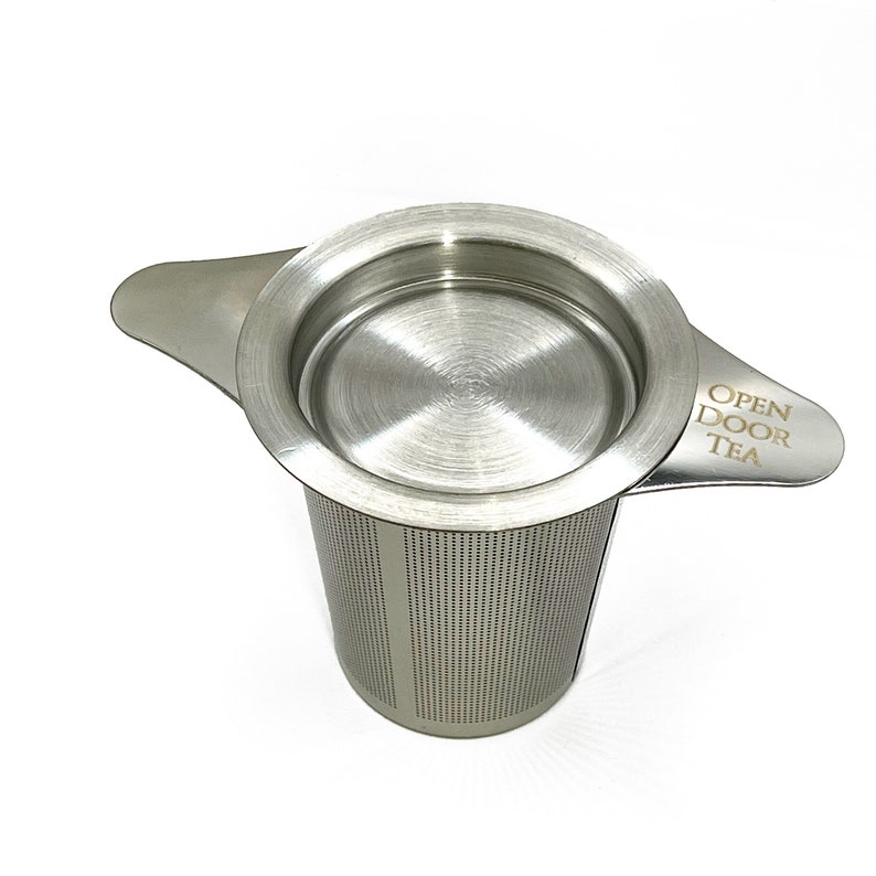 May include: Stainless steel tea infuser with a lid and two handles. The infuser has a fine mesh and the text "OPEN DOOR TEA" is engraved on one of the handles.