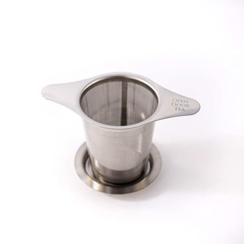May include: A stainless steel tea infuser with a handle and a mesh filter. The infuser has a base that sits on the bottom of a cup or teapot. The infuser has the words "Open Door Tea" printed on the handle.