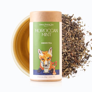 May include: A cylindrical container of Moroccan Mint Green Tea. The label features a fox illustration and the text "Open Door Tea". The tea is loose leaf, with a weight of 2 ounces (56g). A cup of tea is visible on the left, and loose tea leaves are scattered on the right.