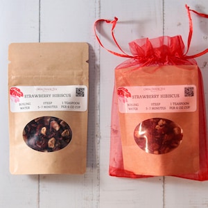 May include: Two packages of loose leaf tea, one in a brown paper pouch and the other in a red mesh bag. Both packages contain dried strawberry hibiscus tea and have a label that reads "OPEN DOOR TEA STRAWBERRY HIBISCUS BOILING WATER STEEP 5-7 MINUTES 1 TEASPOON PER 6 OZ CUP".