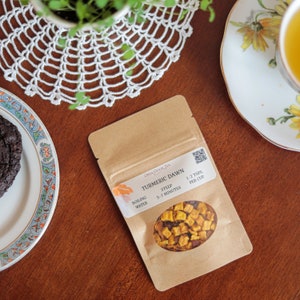 May include: A brown paper pouch with a label that reads "Turmeric Dawn" Open Door Tea. The label also includes instructions for brewing the tea: "Boiling Water, Steep 5-7 Minutes, 1-2 tsps per cup". The pouch contains dried turmeric tea with pieces of dried fruit.