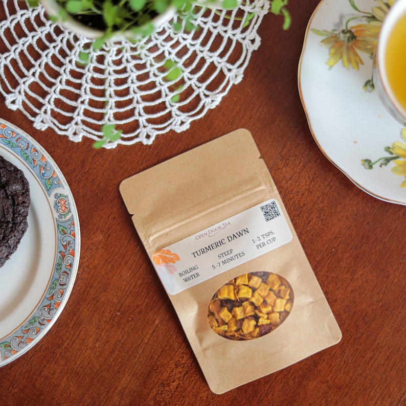 May include: A brown paper pouch with a label that reads "Turmeric Dawn" Open Door Tea. The label also includes instructions for brewing the tea: "Boiling Water, Steep 5-7 Minutes, 1-2 tsps per cup". The pouch contains dried turmeric tea with pieces of dried fruit.