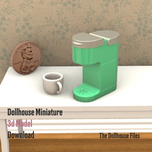 Puede incluir: Una cafetera verde en miniatura con tapa plateada, una taza de café de cerámica blanca y un centavo sobre una mesa blanca. El texto "Dollhouse Miniature 3d Model Download The Dollhouse Files" está impreso en la mesa.