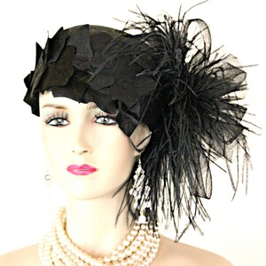 May include: A black hat with a large feather and a black mesh veil. The hat is made of fabric and has a rounded shape. The feather is long and flowing, and the veil is delicate and sheer.