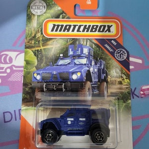 Könnte beinhalten: Ein blauer Matchbox-Spielzeug-LKW aus Metallguss mit schwarzem Kühlergrill, schwarzen Rädern und einem weißen "POLICE"-Aufkleber an der Seite. Der LKW ist auf der Verpackung mit "OSHKOSH DEFENSE M-ATV" und "JUNGLE" beschriftet.