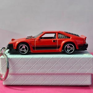May include: A red toy car with black detailing and silver wheels is displayed on a white box. The car is a miniature sports car replica. A silver keyring is attached to the car, suggesting it's a keychain.