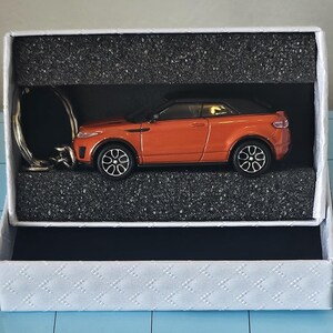 May include: An orange die-cast model car keyring with black wheels and a black roof, presented in a white gift box with a black interior. The keyring is silver. The box has a textured, diamond pattern.