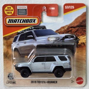 Puede incluir: Un coche de juguete Matchbox Toyota 4Runner 2019 plateado sobre un respaldo de cartón. El embalaje incluye el logotipo de Matchbox, la marca Toyota y el texto "Designed for a Lifetime". El coche de juguete es plateado con ruedas negras.