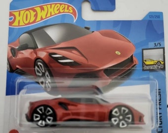 Op de afbeelding: Rode Hot Wheels-speelgoedauto van metaal, die een Lotus Emira voorstelt. De auto staat op een blauwe en witte kaart met het Hot Wheels-logo en de woorden "Lotus Emira". De auto is genummerd 121/250 en 3/5.