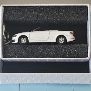 May include: A white model convertible car presented in a white textured gift box with black foam. The car features detailed wheels and a red interior. The box is open, showing the car's side view.