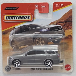 Può includere: Un'auto modello Matchbox Dodge Durango 2018 grigia. La confezione è rossa e gialla con il logo Matchbox e la scritta "2018 Dodge Durango". Il modello è progettato con parti metalliche ed è adatto a bambini dai 3 anni in su.
