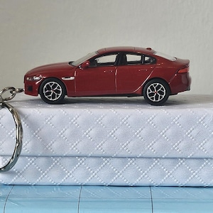 Könnte beinhalten: Ein rotes Modellauto, ca. 10 cm lang, steht auf einer weißen Schachtel mit Steppmuster. Ein silberner Schlüsselanhänger ist am Auto befestigt. Das Auto hat schwarze Räder und einen detaillierten Innenraum.