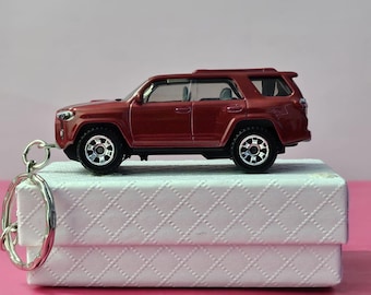 Puede incluir: Un llavero de coche de juguete SUV granate con ruedas negras y un llavero plateado. El coche de juguete está sobre una caja blanca con un patrón de diamantes. El fondo es de color rosa liso.