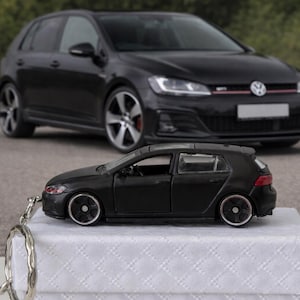 May include: A black toy car keyring, a miniature hatchback replica, rests on a white surface. A full-size black car is in the background. The keyring has silver details and detailed wheels.