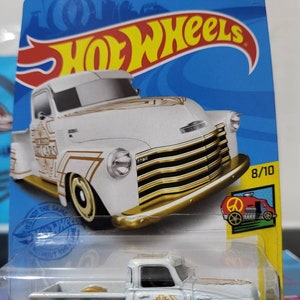 Peut inclure: Un camion Chevrolet blanc de 1949 avec des accents dorés et l'inscription "Art Cars" sur le côté. Le camion est une voiture miniature Hot Wheels en métal moulé sous pression. L'emballage montre le camion sur un fond bleu avec le logo Hot Wheels.