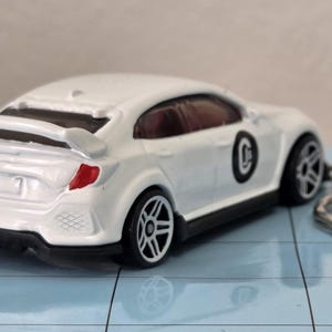 May include: A white toy car with black detailing, a rear spoiler, and a keychain. The car features a red rear light and a black circle with a white "C" on the side. The wheels are silver and black.