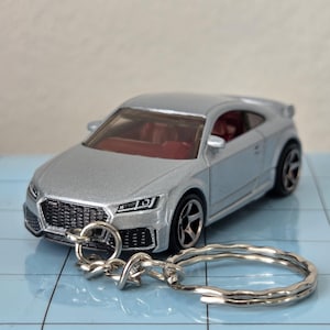 Puede incluir: Coche de modelo plateado con interior rojo, sujeto a un llavero plateado. El coche tiene ruedas negras y una parrilla delantera detallada. El llavero es de metal plateado.